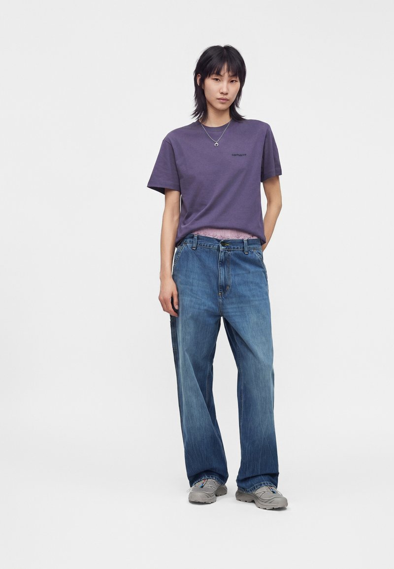 Young person wearing a purple t-shirt, loose blue jeans, gray sneakers, and a necklace, standing with one hand in pocket on white background.