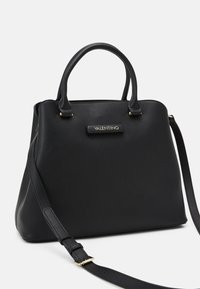 Svart läderhandväska med dubbla handtag och en avtagbar axelrem. Har en guldlogotypplakett på framsidan. Slät textur.