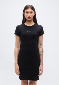 Σύντομο μαύρο φόρεμα από ριμπ ύφασμα, με κοντά μανίκια και στρογγυλή λαιμόκοψη. Ένα λογότυπο βρίσκεται στην περιοχή του στήθους.