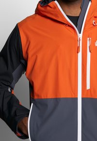 De jas heeft een kleurblokontwerp met een oranje bovenstuk en een grijze onderhelft, gemaakt van een soepel, lichtgewicht materiaal. Witte rits en accenten.