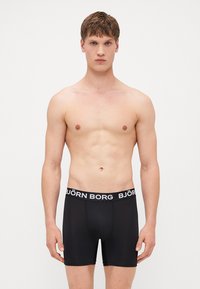 Schwarze Boxer-Shorts mit breitem Gummibund, auf dem der Text "BJÖRN BORG" in Weiß zu sehen ist. Glattes, dehnbares Material. Model steht, Oberkörper sichtbar.