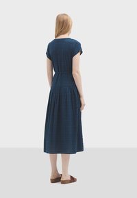 Dunkelblaues, kurzärmeliges Kleid mit strukturiertem Muster, tailliertem Schnitt und fließendem Rock. Getragen mit braunen Schlüpfschuhen.