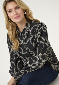 Camicia nera con bottoni, caratterizzata da un motivo a catena bianco, maniche lunghe con polsini elastici e una vestibilità rilassata. Tessuto dalla consistenza liscia.