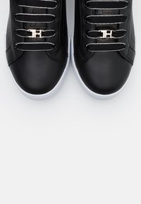 Ein Paar schwarze Leder-Sneaker mit weißen Sohlen, schwarzen Schnürsenkeln mit weißen Rändern und goldfarbenen Schnürsenkelanhängern, die mit den Initialen "TH" geprägt sind.