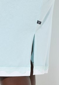 Lyseblå bomuldsskjørt med sideslids, hvid bølget kant og et sort mærke i siden. Blød tekstur og afslappet design.