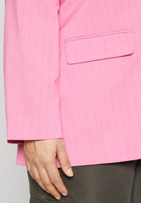 Růžový blazer s vertikálními proužky, klopovým límcem a jednou kapsou na přední straně. Hladká textura s mírně volným střihem.