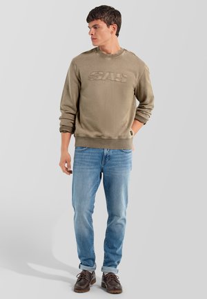 Joven con sudadera marrón con las mangas remangadas, vaqueros azules y zapatos marrones, de pie con una mano en el bolsillo mirando hacia la izquierda.