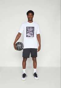 T-shirt blanche à manches courtes avec un graphique de basketball noir, assortie à un short noir et des baskets blanches, tenant un ballon de basketball.
