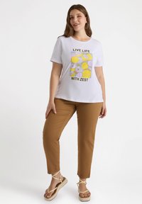 Camiseta blanca con diseño gráfico de limón y lima, texto negro "VIVE LA VIDA CON ENERGÍA", combinada con pantalones marrones y sandalias de tiras beige.