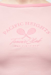 Ροζ υφασμάτινο ύφασμα με ριγέ υφή και κέντημα δύο σταυρωμένων ρακέτων τένις και κείμενο που γράφει "Pacific Heights Tennis Club San Francisco."