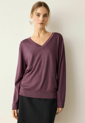 Ung kvinde iført en lilla V-hals sweater og sort nederdel står mod en ensartet lys baggrund med store sølvfarvede hoop-øreringe.