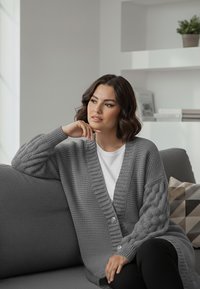 Szary dziergany kardigan z szerokim prążkowanym kołnierzem, detalami na guziki i teksturowanymi rękawami w warkoczowy splot, noszony na białej bluzce i czarnych spodniach.