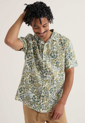 Mann mit lockigem Haar, der ein kurzärmliges Hemd mit blau-grünen Blattmustern trägt, lächelt und sich an den Haaren berührt.