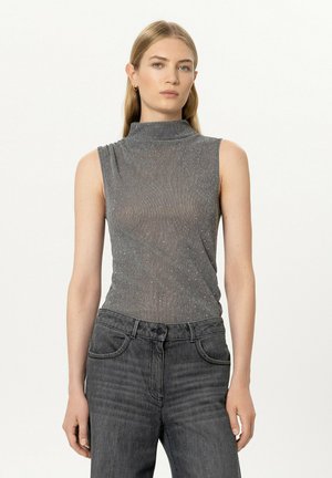 Young woman with long blond hair wearing a sleeveless gray shimmer top and high-waisted dark gray jeans, standing against a plain background.