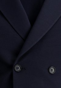 Blazer azul marino de doble botonadura con un tejido texturizado, solapas con muesca y dos botones negros de cuatro orificios en la parte delantera.