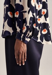 Blus med blommönster i marinblått, cream och orange; har en samlad ärm med knappstängning. Buren över mörkt tyg.