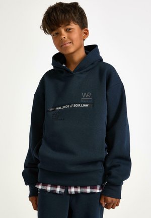 Navy blue hoodie with a soft texture, featuring a front graphic with 'WR' logo and city names. Layered over a plaid shirt.
