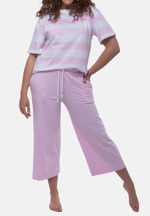 Kobieta ubrana w luźne różowe spodnie o szerokich nogawkach i krótki różowo-biały pasiasty top, stojąca boso z ręką w kieszeni.