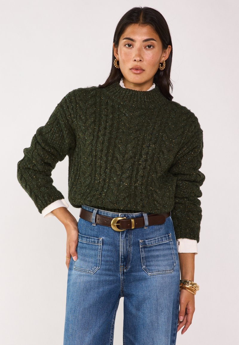 Maglione verde chunky a maglia con texture a coste e colletto alto, sovrapposto a una camicia bianca. Abbinato a jeans blu a vita alta e una cintura marrone.