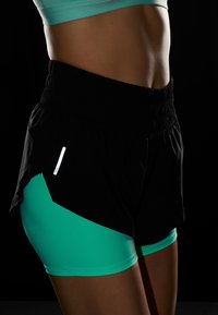 Svarta träningsshorts med elastisk midja, med en teal underlayer och en reflekterande detalj på sidan. Slät textur och åtsittande design.