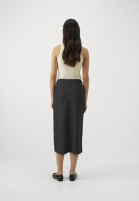 Gonna nera midi con una texture liscia, indossata sopra un top aderente color crema. La gonna ha un design semplice ed è lunga fino alla caviglia. Scarpe nere basse completano l'outfit.