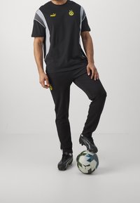 Man klädd i svart Puma Borussia Dortmund fotbollsklädsel står med foten på en Puma fotboll mot en enhetlig bakgrund.