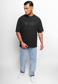 Camiseta negra de manga corta con el texto "Dropsize" en relieve, combinada con unos vaqueros de corte holgado en azul claro y zapatillas blancas. Textura suave.