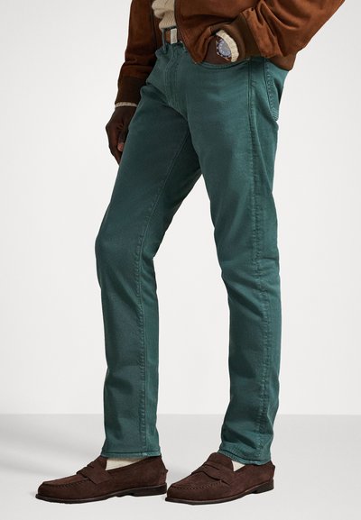 Tirkīza krāsas kokvilnas auduma bikses ar šauru piegriezumu, ar sānu kabatām un minimālu šuvju detaļu. Valkā ar brūniem zamšāda mocasin apaviem.