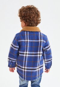 Niebiesko-brązowa koszula flanelowa w kratę z beżowym, sztucznym kołnierzem z owczej wełny, z długimi rękawami i guzikami przy mankietach, widok z tyłu.