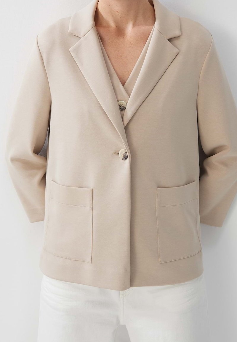 Personne portant un blazer beige avec de grandes poches à l'avant et un pantalon blanc, posant devant un fond uni et clair. Buste visible, mains derrière le dos.