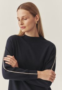 Marineblaues Sweatshirt mit hohem Kragen und langen Ärmeln, das einen kontrastierenden hellen Besatz entlang der Ärmel aufweist. Weiche Textur erkennbar.