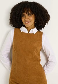Vestido de pana marrón con textura acanalada y tirantes anchos, usado sobre una camisa blanca con cuello. La modelo posa con las manos en las caderas y sonríe.