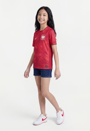 Fille souriante, vêtue d'un maillot de football rouge de la Pologne, d'un short bleu et de baskets blanches Nike avec des virgules rouges, debout sur un fond blanc.