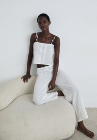 Top alb croppat cu bretele ajustabile și detalii brodate, asortat cu pantaloni albi cu croială relaxată, amândouă având modele texturate subtile.