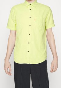 Koszulka z krótkim rękawem w jasnym limonkowym kolorze, z teksturowanej tkaniny, z kołnierzykiem zapinanym na guziki, kieszonką na piersi i ciemnymi guzikami.