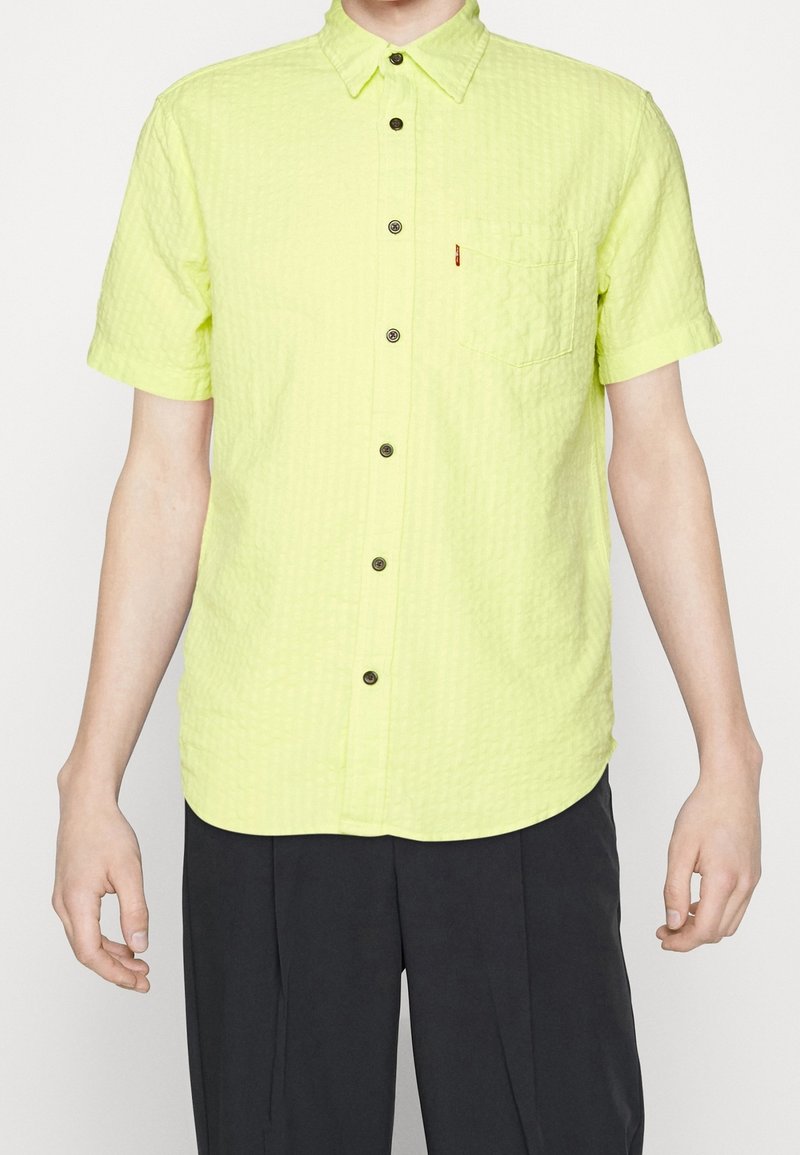 Koszulka z krótkim rękawem w jasnym limonkowym kolorze, z teksturowanej tkaniny, z kołnierzykiem zapinanym na guziki, kieszonką na piersi i ciemnymi guzikami.