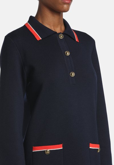 Tmavomodrá dlhá polokošeľa s červeným a bielym lemovaním goliera a vreciek, s ozdobnými zlatými gombíkmi vpredu aj na vreckách.