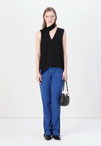 Schwarzes ärmelloses Oberteil mit V-Ausschnitt und Faltenoptik, kombiniert mit blauen weit geschnittenen Hosen. Das Modell hält eine kleine schwarze Handtasche.