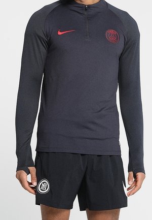 Hombre con camiseta de entrenamiento de manga larga oscura de Paris Saint-Germain Nike y pantalones cortos negros con logotipos blancos, de pie sobre un fondo liso.