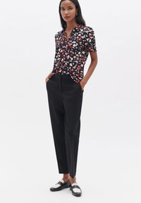 Schwarze Bluse mit floralem Muster und kurzen Ärmeln, rot-weiße Muster; kombiniert mit schwarzen, hochgeschnittenen Hosen und schwarz-weißen Schuhen.