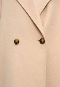 Μπεζ παλτό από μαλλί με διπλό πέτο γιακά, διαθέτει δύο σκούρα καφέ κουμπιά και μαλακή, υφή επιφάνεια.