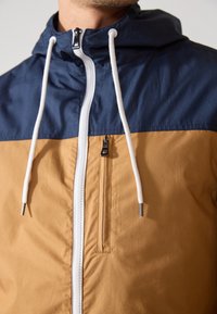 Flerfärgad jacka med en marinblå övre del och en ljusbrun nedre del. Har dragkedja framtill, justerbar huva och vita dragsnören.