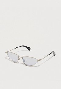 Óculos de sol com armação prateada, hastes pretas finas e lentes angulares em formato de gato ligeiramente coloridas, sobre uma superfície branca.