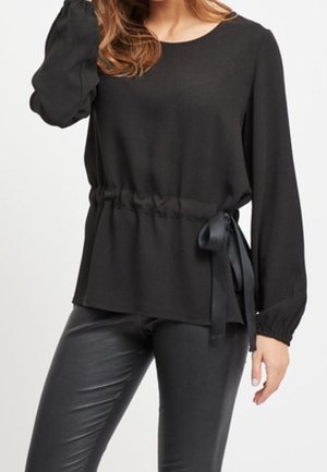 Kvinde iført en sort langærmet bluse med indsnævring ved taljen og bånd som bindebånd, kombineret med sorte bukser i læderlook.