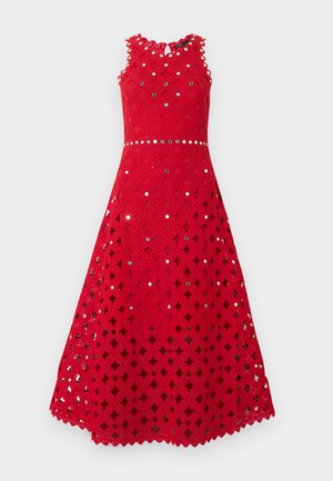 Frau trägt ein ärmelloses rotes Midikleid mit Lochspitzenmuster und weiße spitze Schuhe, steht vor einem einfarbigen hellen Hintergrund.