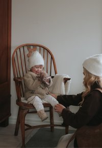 Lapsi, joka on pukeutunut beigeen pörröiseen takkiin ja valkoiseen hattuunsa, istuu puisella tuolilla lelua pitelemässä. Pehmeä valkoinen viltti peittää tuolia; ruskeaan pukeutunut nainen on vuorovaikutuksessa lapsen kanssa.