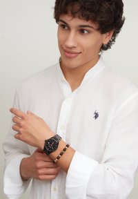 Orologio da polso nero con quadrante ottagonale e bracciale in argento indossato su un polso, abbinato a un braccialetto di perline. Il modello indossa una camicia di lino bianca.