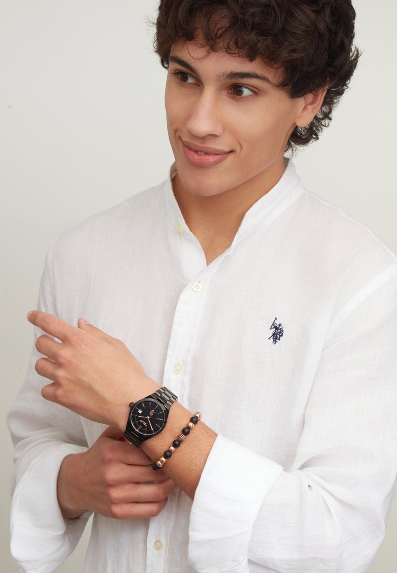 Orologio da polso nero con quadrante ottagonale e bracciale in argento indossato su un polso, abbinato a un braccialetto di perline. Il modello indossa una camicia di lino bianca.