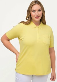 Koszulka polo z krótkim rękawem w kolorze żółtym, wykonana z miękkiego materiału, z kołnierzykiem, listwą guzikową oraz kieszenią na piersi. Gładka tekstura z luźnym krojem.