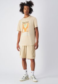 Camiseta oversized beige con gráfico de "Demasiado Caliente"; pantalones cortos cargo a juego; calcetines blancos; zapatillas color crema con acentos negros.
